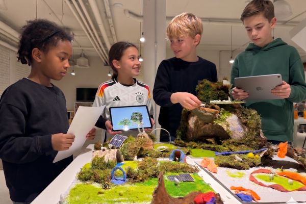 Kinder mit iPads und Computern stehen vor einem Tisch voller Modelle